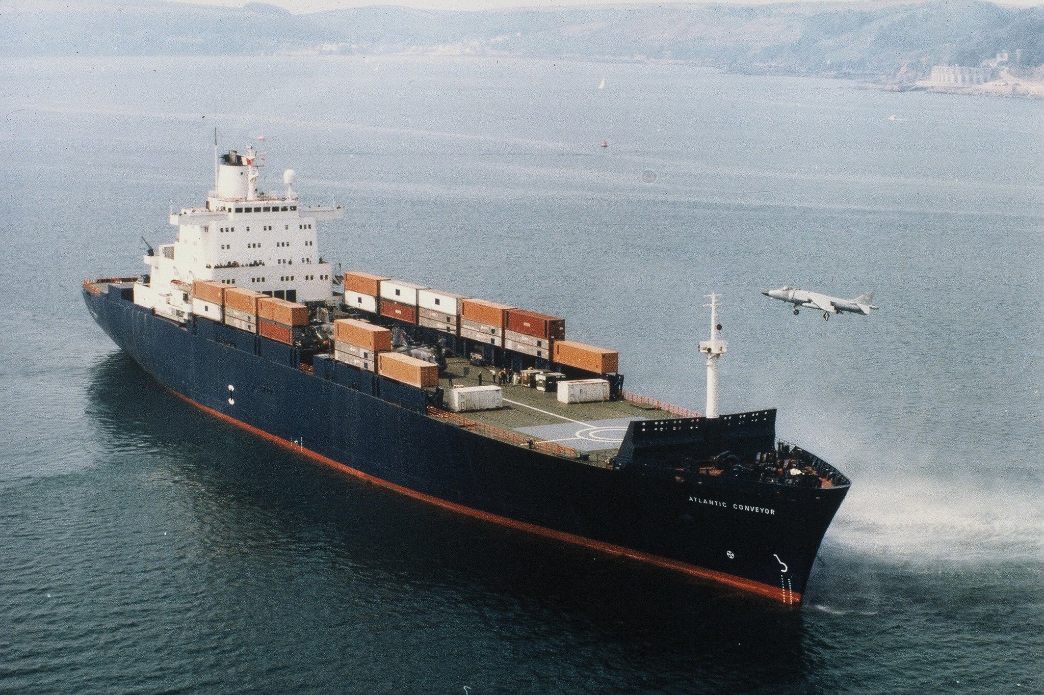 Aerial starboard bow view of the British Merchant Ship Atlantic Conveyor with a Harrier jump jet hovering near the bow.