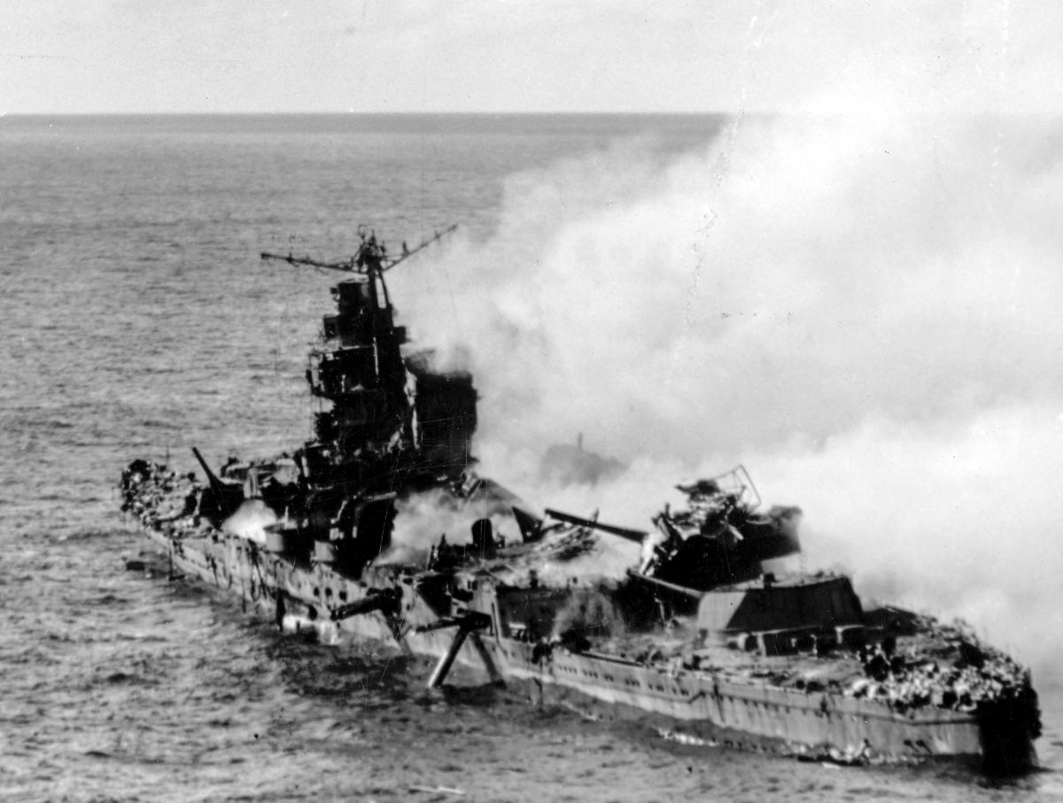 The Japanese heavy cruiser, Mikuma, smolders off Midway, the wreckage of a Marine dive bomber on one of turrets.