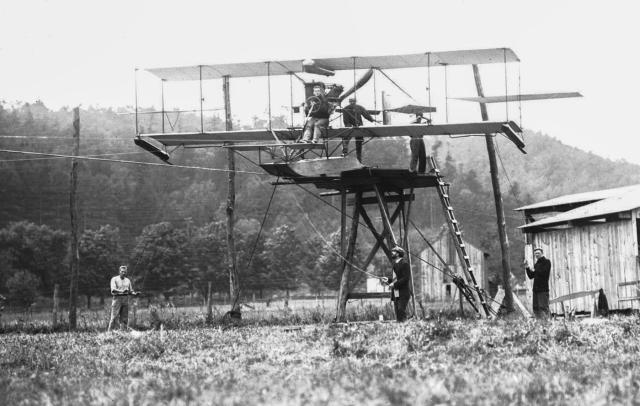 Lieutenant Theodore Ellyson is in the pilot’s seat of A-1 as preparations are made for the cable test launch. The main cable ran in an inverted U-channel beneath the pontoon; thinner cables were strung near each wingtip for balance. The broad U-shaped tip guides are visible beneath the wings.