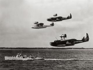 A cutter is passed overhead by three flying life boats. Capable of at-sea rescues, the FLBs (or PJs) were the first aircraft designed from the start for Coast Guard service.
