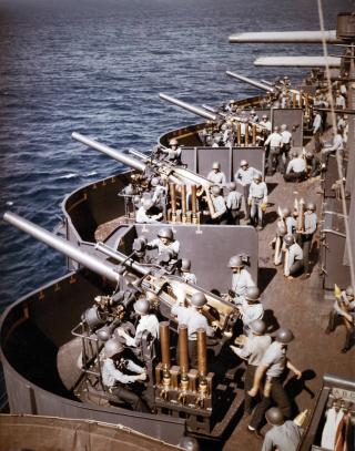 The 5-inch/25-caliber battery on board the USS North Carolina (BB-40) prepares to fire during the bombardment of Saipan, 15 June 1944. Note the time-fuze setters on the left side of each gun mount, each holding three fixed rounds of ammunition; dark barrels of 20-mm guns at the extreme right; and triple 14/50 guns in the background.