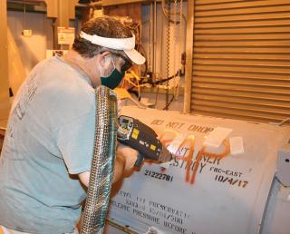 A Fleet Readiness Center East worker demonstrates using laser ablation to remove a swath of coating from an aircraft engine container. Laser ablation employs a noncontact technique to remove material, eliminating the need for mechanical tools and significantly reducing the amount of noise and waste produced.