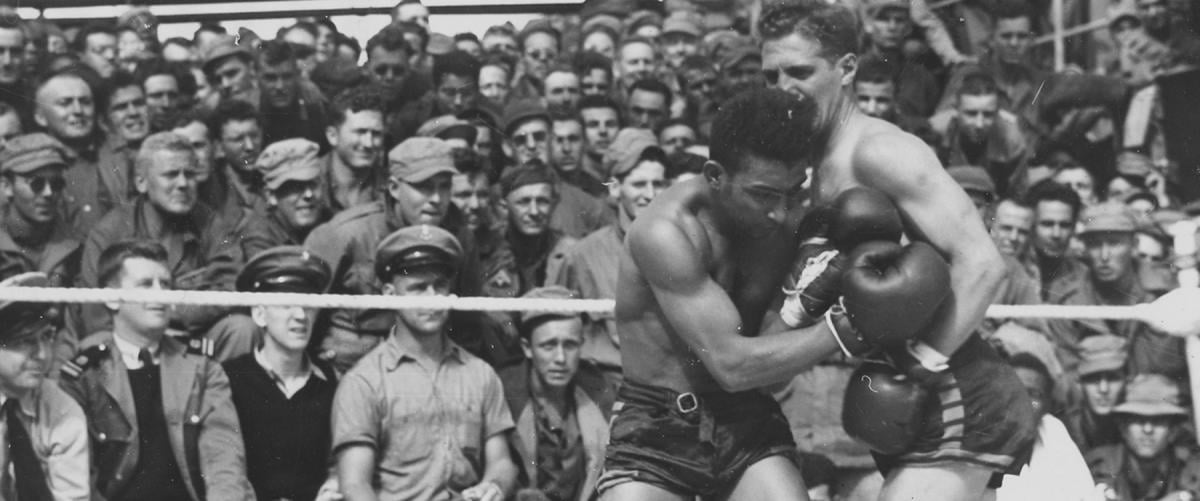 Retired professional boxer and Coast Guard Commander Jack Dempsey (left) gives an added punch when he referees a boxing match on the deck of a Coast Guard-manned troop transport headed for Europe during World War II.