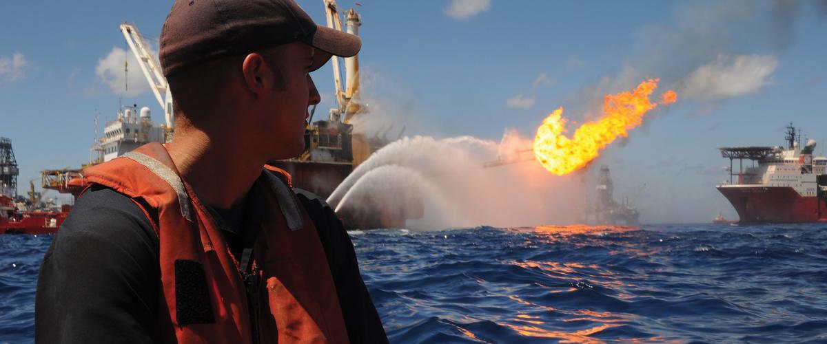 A Coast Guard petty officer looks on from a small boat detached from the USCGC Resolute (WMEC-620)  as oil and gas are flared off at the site of the 2010 Deepwater Horizon oil spill. A post-incident review identified  gaps in the Coast Guard’s marine environmental response preparedness and programs.