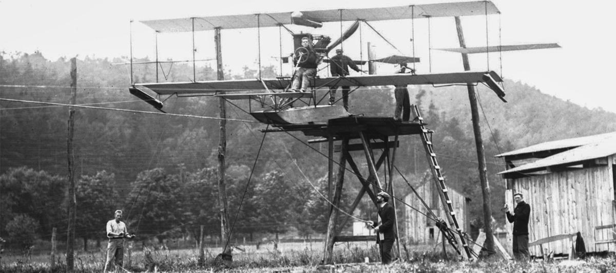 Lieutenant Theodore Ellyson is in the pilot’s seat of A-1 as preparations are made for the cable test launch. The main cable ran in an inverted U-channel beneath the pontoon; thinner cables were strung near each wingtip for balance. The broad U-shaped tip guides are visible beneath the wings.