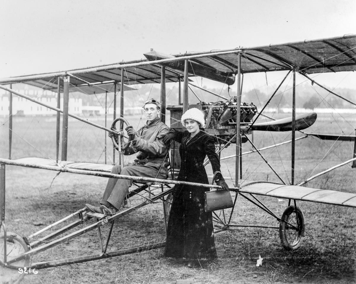  Eugene B. Ely seated in a Curtiss pusher biplane, with his wife, Mabel (Hall) Ely standing beside him