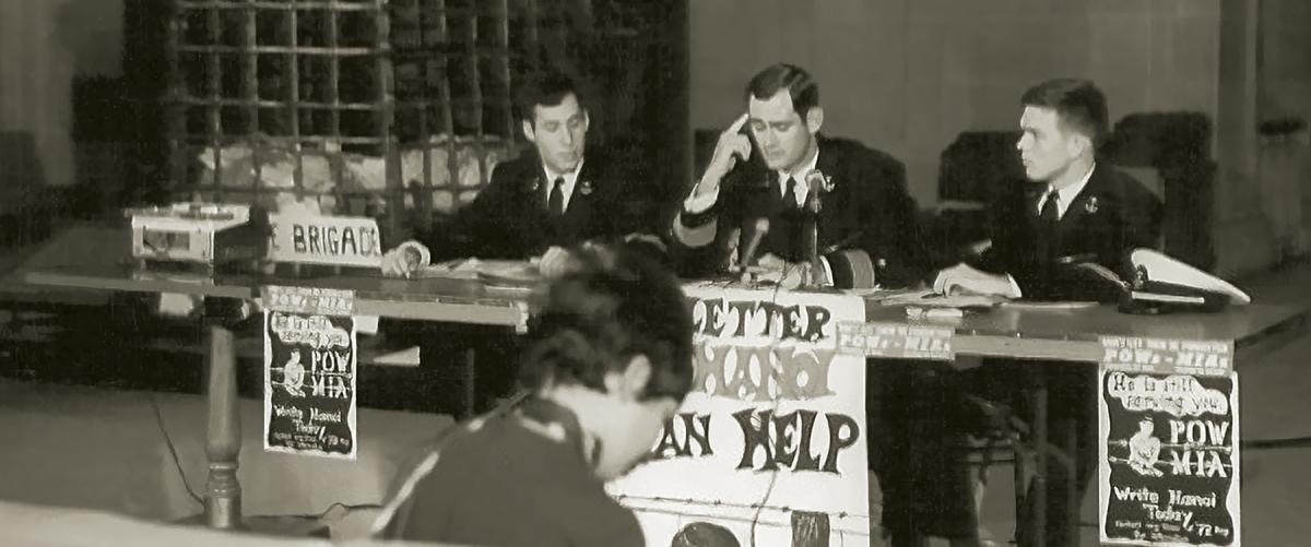 In fall 1970, the Naval Academy held a press conference on the plight of POW/MIA families. Midshipmen Rick Rubel and Joe Glover flank Brigade Commander Mike Hecomovich, with the replica bamboo cage in the background.