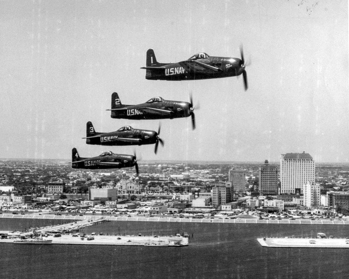 An aerial right side view of four F8F-1 Bearcat fighter aircraft in formation over Corpus Christi's water front area.