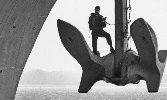 A sailor chips paint from his ship’s anchor while at Naval Air Station Pensacola. Chipping paint and repainting the anchor is an important part of regular ship maintenance. Circa 1982.