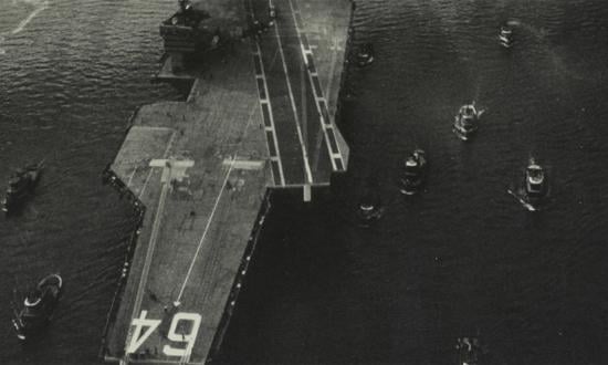 Aerial view of the USS Constellation (CVA-64) surrounded by harbor tugs as it passes beneath the Manhattan Bridge in January 1962. Brooklyn Bridge is visible in the background.