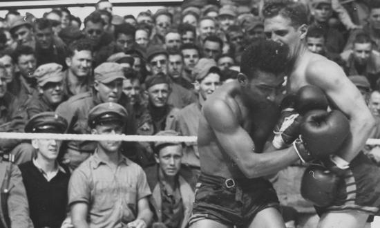 Retired professional boxer and Coast Guard Commander Jack Dempsey (left) gives an added punch when he referees a boxing match on the deck of a Coast Guard-manned troop transport headed for Europe during World War II.