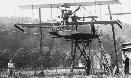 Lieutenant Theodore Ellyson is in the pilot’s seat of A-1 as preparations are made for the cable test launch. The main cable ran in an inverted U-channel beneath the pontoon; thinner cables were strung near each wingtip for balance. The broad U-shaped tip guides are visible beneath the wings.