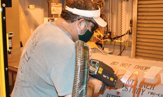 A Fleet Readiness Center East worker demonstrates using laser ablation to remove a swath of coating from an aircraft engine container. Laser ablation employs a noncontact technique to remove material, eliminating the need for mechanical tools and significantly reducing the amount of noise and waste produced.