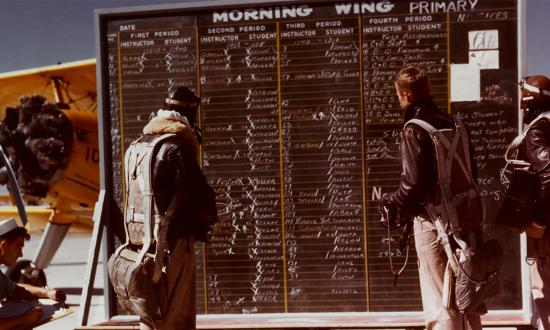 Flight instructors and naval aviation cadets check the operations board for the morning flight schedule, at a naval air station, circa 1942-1943. Note Stearman N2S parked behind the board.  Credit: National Archives